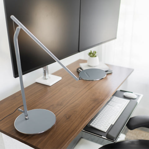 Top view of a office desk with a keyboard tray, desk lamp, double monitors and a plant.